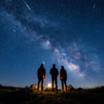 Trois silhouettes observent un ciel nocturne rempli d'étoiles, de la Voie Lactée et de nombreuses étoiles filantes.