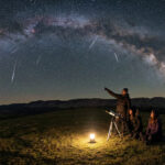 Trois personnes observent le ciel étoilé avec un télescope sous un arc spectaculaire de la Voie lactée.