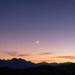 Un croissant de Lune brillant au-dessus d'une chaîne de montagnes lors d'un crépuscule aux nuances pourpres.