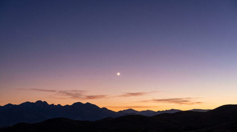 Un croissant de Lune brillant au-dessus d'une chaîne de montagnes lors d'un crépuscule aux nuances pourpres.